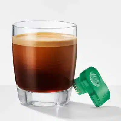Un verre rempli d'une boisson au café en couches, doté d'un dessus mousseux. Attaché au bord se trouve Payment Connect, un petit appareil électronique vert avec des connecteurs visibles, indiquant qu'il pourrait être utilisé pour la transmission de données ou la détection liée à la boisson.