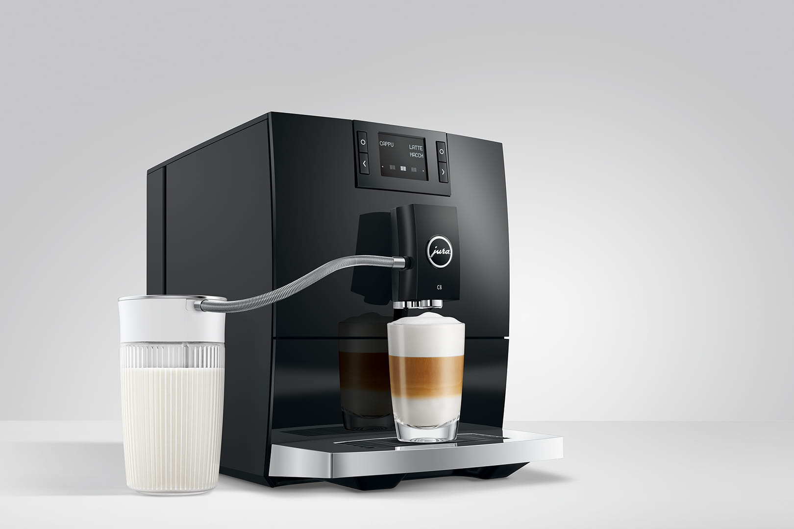 Un JURA C8 Piano Black (SA) avec commandes numériques prépare un latte. La machine dispose d'un tube pour faire mousser le lait relié à un verre de lait sur le côté. Un latte avec plusieurs couches de lait et de café est distribué dans une tasse en verre transparent posée sur le plateau.