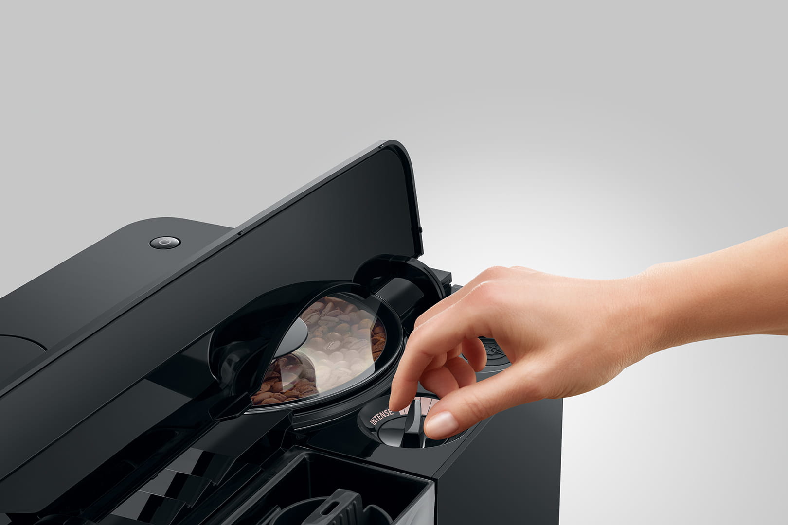 Gros plan de la main d'une personne ajustant un réglage sur une machine à café JURA C8 Piano Black (SA). La machine dispose d'un compartiment contenant des grains de café et de divers cadrans et boutons pour la personnalisation. Le fond est uni et gris.