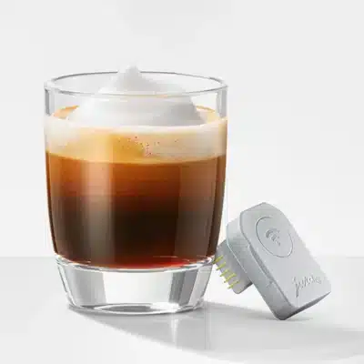 Un verre transparent rempli d'une boisson au café en couches, avec de la mousse de lait mousseuse sur le dessus. À côté du verre se trouve un petit appareil électronique gris appelé MDB Connect, avec un symbole Wi-Fi et des broches de connexion. Le fond est simple et blanc.