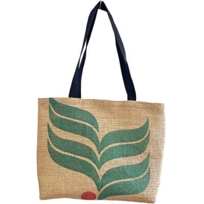 Eine Jute-Tragetasche mit schwarzen Griffen, einem stilisierten grünen Blattdesign und einem kleinen roten Kreis am unteren Rand. Die natürliche Farbe des Jutegewebes kontrastiert mit dem lebendigen Druck.