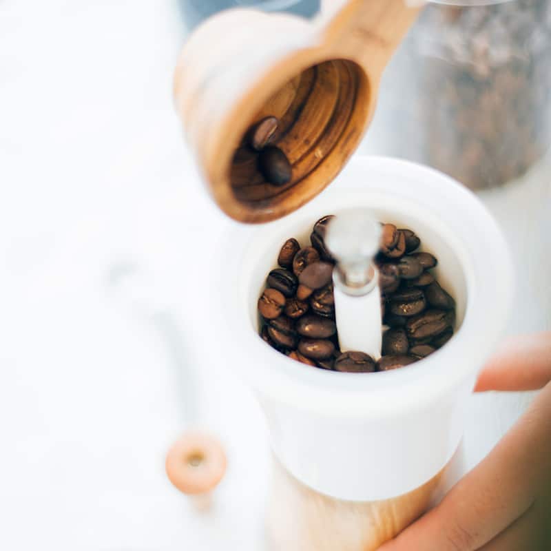 Gros plan des mains manipulant le HARIO - Moulin à café manuel en céramique et olivier rempli de grains de café. Ce moulin est doté d'un corps en céramique blanche et d'un manche en bois. L'arrière-plan est flou, dévoilant subtilement un autre moulin et un récipient.