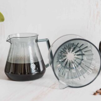 Une installation de café avec le dripper TIME MORE - Crystal Eye inversé avec du café fraîchement moulu dans une carafe en verre, à côté d'un moulin à café manuel, le tout placé sur une surface claire. Une plante en pot est visible en arrière-plan.