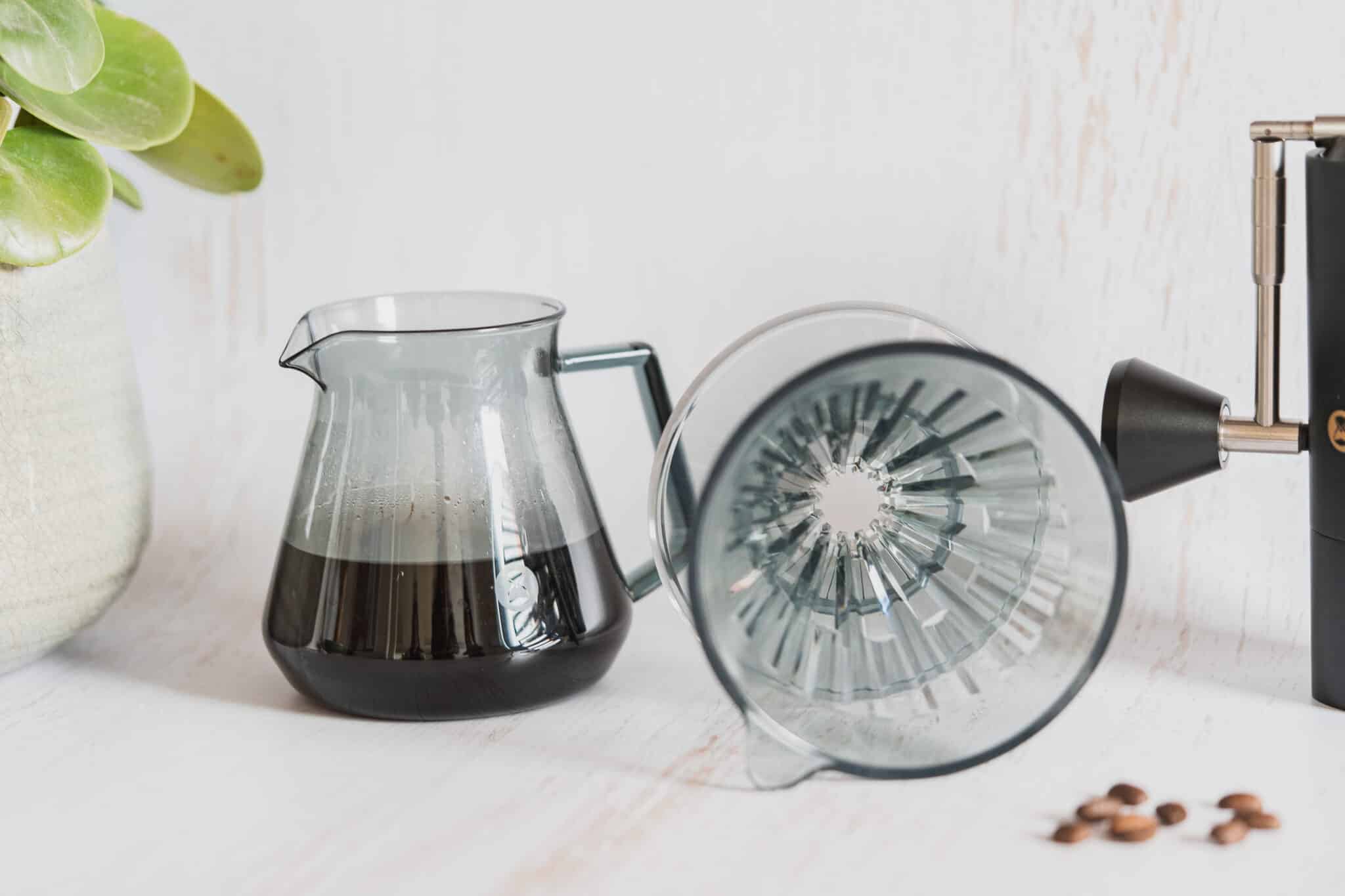 Une installation de café avec le dripper TIME MORE - Crystal Eye inversé avec du café fraîchement moulu dans une carafe en verre, à côté d'un moulin à café manuel, le tout placé sur une surface claire. Une plante en pot est visible en arrière-plan.