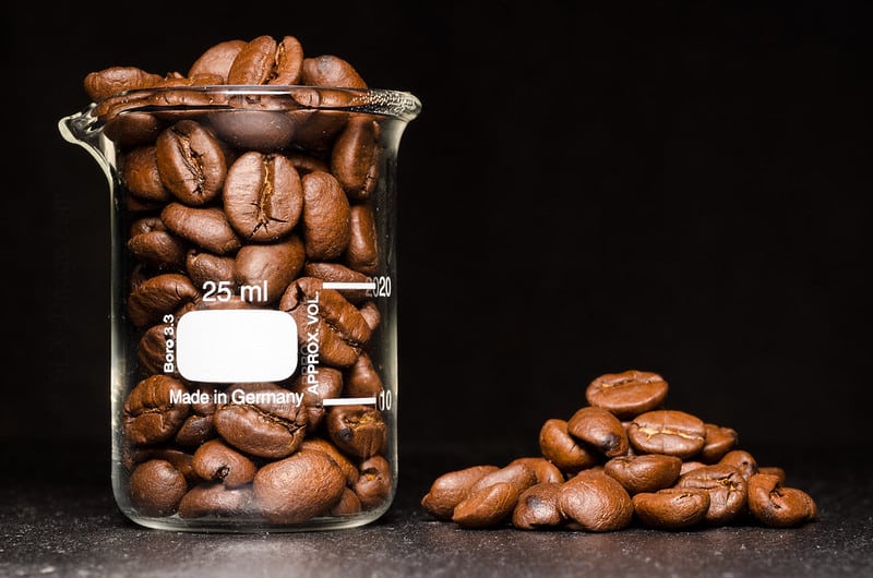 Un récipient en verre de 25 ml rempli de grains de café torréfiés repose sur une surface sombre, évoquant le riche arôme des boissons à base de café. D'autres grains de café sont dispersés à droite du récipient sur un fond noir, renforçant cette scène captivante.