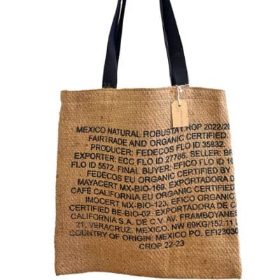 Eine Einkaufstasche aus Jute mit aufgedrucktem Text, der Angaben zu Fairtrade- und Bio-Zertifizierung, Produzent, Exporteur und Anbauinformationen für Kaffee enthält. Die Tasche hat schwarze Griffe und ein befestigtes Etikett.