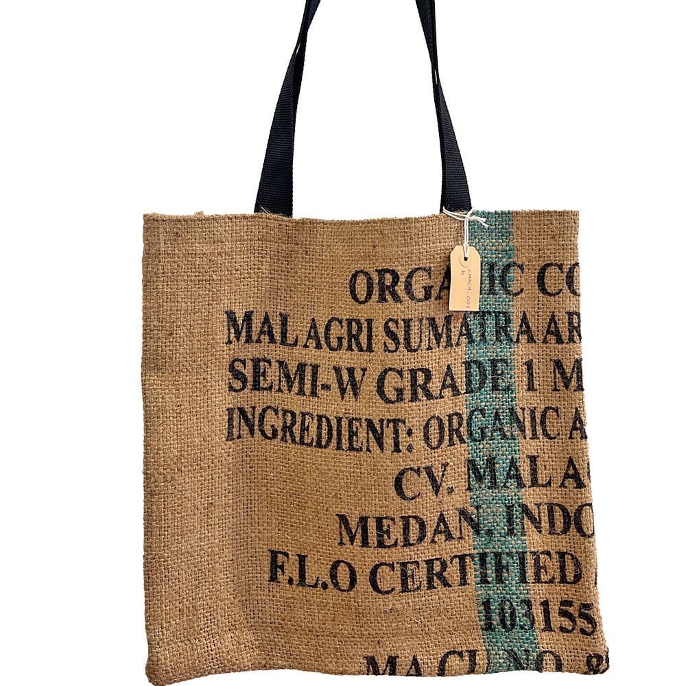 Eine Einkaufstasche aus Jute mit schwarzem Text und Angaben zum Kaffee wie Herkunft und Zertifizierung. Sie hat zwei dunkle Griffe und ein kleines Etikett.
