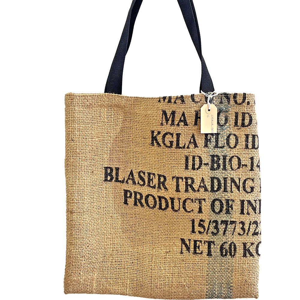 Eine Einkaufstasche aus Jute mit dunklen Griffen, aufgedrucktem Text wie „Blaser Trading“ und „Product of India“. An einem Griff ist ein kleines beiges Etikett befestigt.